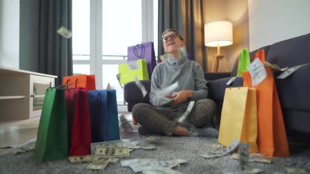 Boldog nő ül a szőnyegen egy hangulatos szobában között bevásárló táskák és pénzt eső amerikai dollár számlák — Stock videók