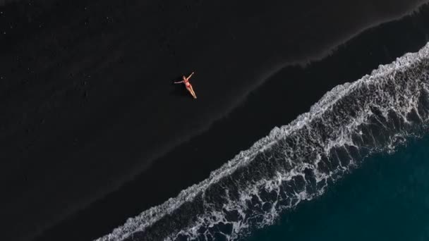 Bovenaanzicht van een meisje in een rood badpak liggend op een zwart strand op de surflijn. Kust van het eiland Tenerife, Canarische Eilanden, Spanje. — Stockvideo