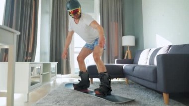 Eğlenceli video. Şortlu ve tişörtlü bir adam rahat bir odada halının üzerinde snowboard yaparken resmediyor. Karlı bir kış bekliyor