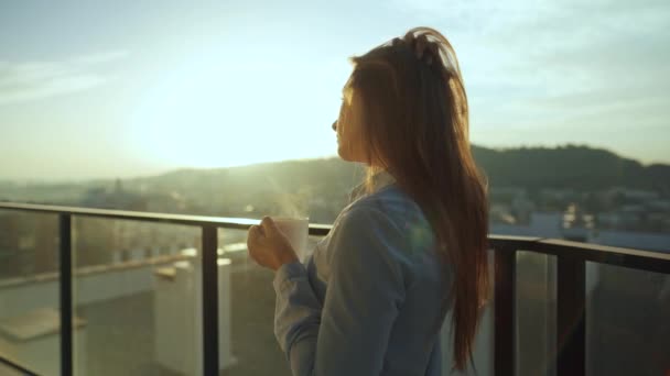 Hübsche Frau trinkt eine Tasse Kaffee oder Tee, beobachtet einen wunderschönen Blick auf die Stadt und genießt entspannt die frische frierende Luft auf dem Balkon bei Sonnenaufgang — Stockvideo