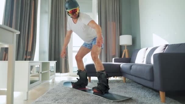 Un video divertente. Uomo in pantaloncini e una T-shirt raffigura snowboard su un tappeto in una stanza accogliente. Aspettando un inverno nevoso — Video Stock