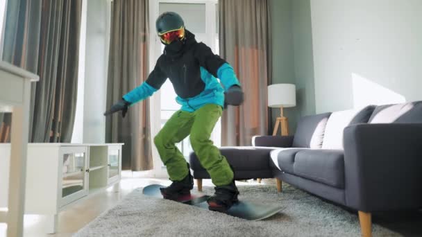 Vídeo divertido. Hombre vestido de snowboarder monta una tabla de snowboard en una alfombra en una habitación acogedora. Esperando un invierno nevado — Vídeos de Stock