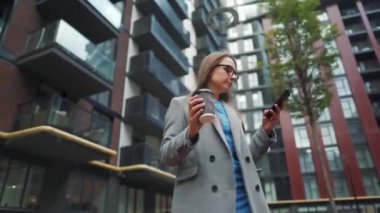 Resmi olarak giyinmiş bir kadın bir iş bölgesinde elinde kahve ve akıllı telefon kullanarak yürüyor.