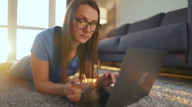 Gözlüklü kadın yerde yatıyor ve kredi kartı ve dizüstü bilgisayar kullanarak online alışveriş yapıyor. İnternetten alışveriş, yaşam tarzı teknolojisi.