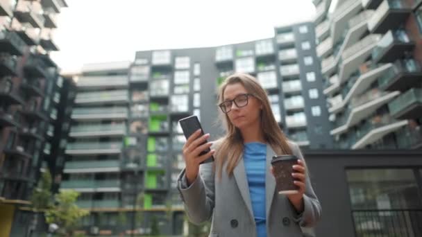 Donna formalmente vestita che cammina per strada in un quartiere degli affari con il caffè in mano e utilizzando uno smartphone — Video Stock