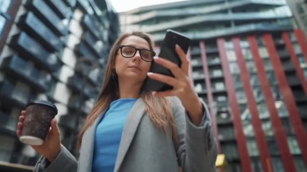 Mulher formalmente vestida de pé em um distrito de negócios com café na mão e usando um smartphone. Câmera se move em torno dela — Vídeo de Stock