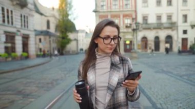Elinde gözlüklü, ceketli ve termosu olan bir kadın eski bir caddede yürüyor ve akıllı telefon kullanıyor..