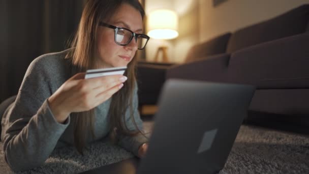眼鏡をかけた女性が床に横になっており、夕方にクレジットカードやノートパソコンを使ってオンラインで購入しています。オンラインショッピング、ライフスタイル技術 — ストック動画