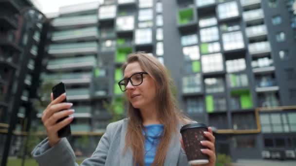 Mujer vestida formalmente caminando por la calle en un distrito de negocios con café en la mano y hablando en una videollamada para teléfonos inteligentes — Vídeos de Stock