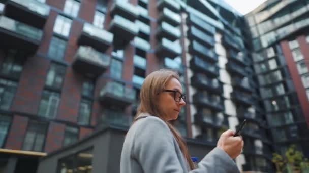 Mujer vestida formalmente de pie en un distrito de negocios con la taza termo en la mano y utilizando un teléfono inteligente. Cámara se mueve alrededor de ella — Vídeo de stock