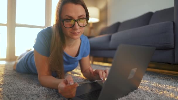 Kobieta w okularach leży na podłodze i dokonuje zakupu online za pomocą karty kredytowej i laptopa. Zakupy online, technologia stylu życia — Wideo stockowe