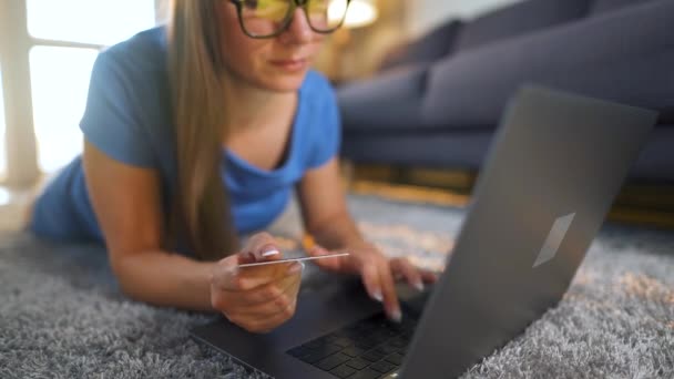 Silmälasipäinen nainen makaa lattialla ja tekee verkko-ostoksen luottokortilla ja kannettavalla tietokoneella. Verkkokaupoissa, elämäntapa tekniikka — kuvapankkivideo