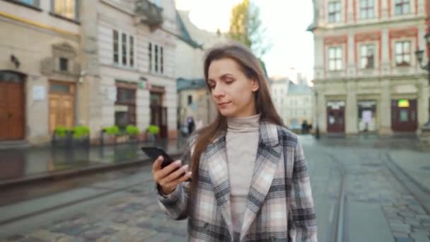 Mujer vistiendo un abrigo caminando por una calle vieja y usando un teléfono inteligente — Vídeo de stock