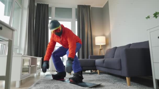 Vídeo divertido. Hombre vestido de snowboarder monta una tabla de snowboard en una alfombra en una habitación acogedora. Esperando un invierno nevado — Vídeos de Stock