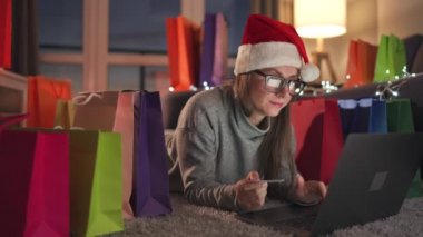 Noel Baba şapkası takan mutlu kadın halıda yatıyor ve kredi kartı ve dizüstü bilgisayar kullanarak online alışveriş yapıyor. Alışveriş torbaları..