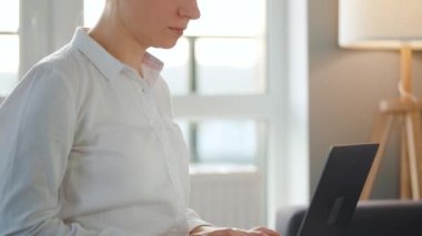 Kadın sıcak bir odada kanepede oturuyor ve gün batımında dizüstü bilgisayarla çalışıyor. Uzak çalışma kavramı.