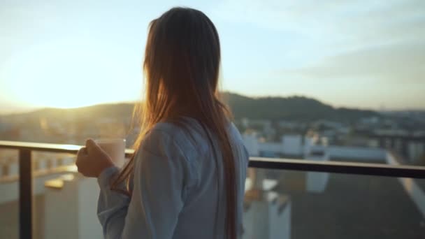 Bella donna beve una tazza di caffè o tè, guardando una bella vista urbana e godersi il relax respirando aria fresca congelamento sul balcone all'alba — Video Stock