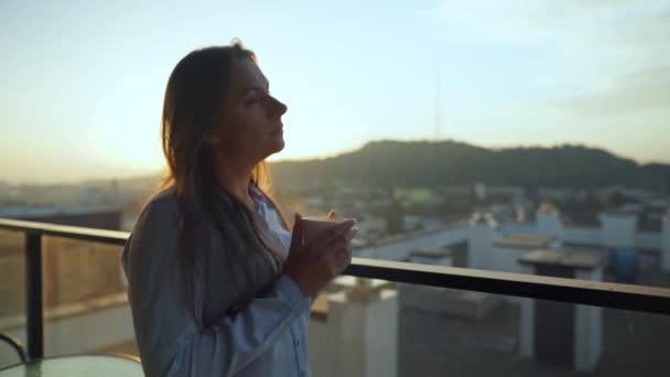 Hübsche Frau trinkt eine Tasse Kaffee oder Tee, beobachtet einen wunderschönen Blick auf die Stadt und genießt entspannt die frische frierende Luft auf dem Balkon bei Sonnenaufgang — Stockvideo