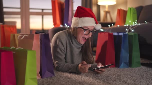 Mujer feliz con gafas con un sombrero de Santa Claus está acostado en la alfombra y hace una compra en línea con una tarjeta de crédito y un teléfono inteligente. Bolsas de compras alrededor. — Vídeo de stock