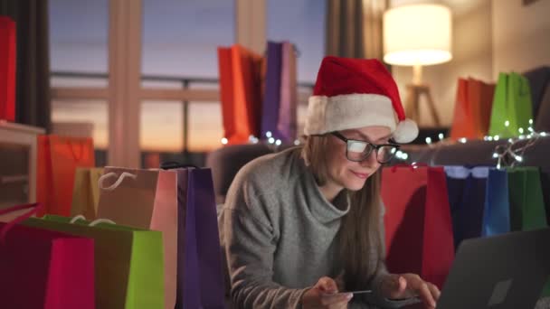 Mujer feliz con gafas con un sombrero de Santa Claus está acostado en la alfombra y hace una compra en línea con una tarjeta de crédito y un ordenador portátil. Bolsas de compras alrededor. — Vídeo de stock