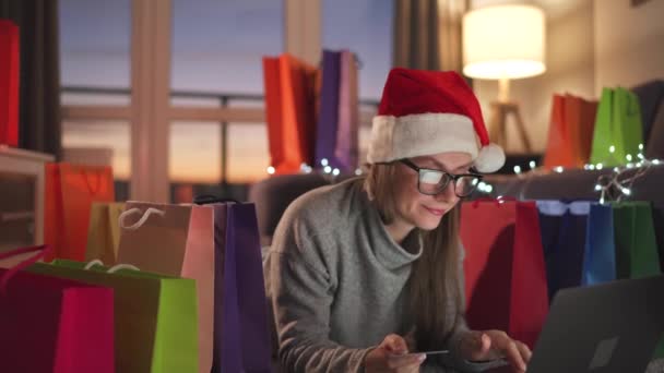 Mujer feliz con gafas con un sombrero de Santa Claus está acostado en la alfombra y hace una compra en línea con una tarjeta de crédito y un ordenador portátil. Bolsas de compras alrededor. — Vídeos de Stock