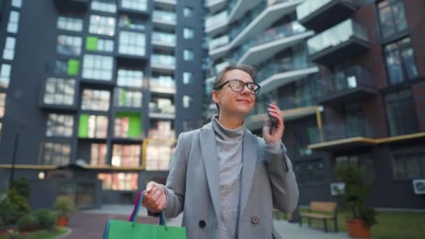 Mulher sorridente andando em um distrito de negócios com sacos de compras e falando no smartphone. Estilo de vida urbano e conceito de tecnologia digital — Vídeo de Stock