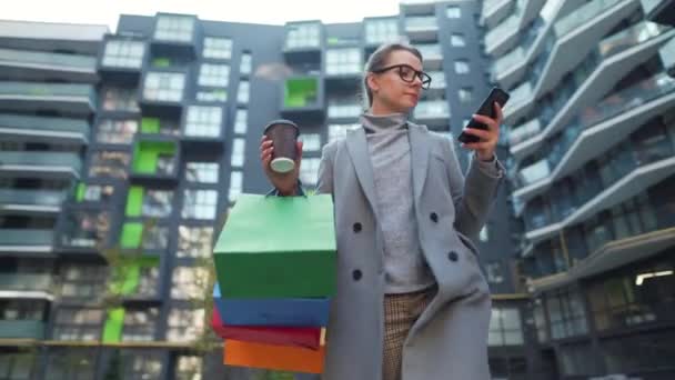 Mujer caminando en un distrito de negocios, sostiene el café para llevar, bolsas de compras y utiliza el teléfono inteligente. Estilo de vida urbano y concepto de tecnología digital — Vídeo de stock