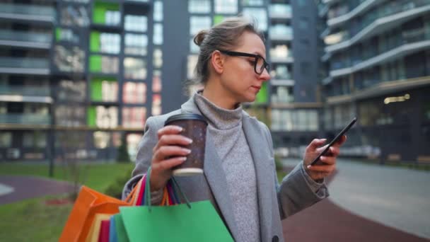 Frau steht im Geschäftsviertel, hält Kaffee zum Mitnehmen, Einkaufstüten und nutzt Smartphone. Urbaner Lebensstil und digitales Technologiekonzept — Stockvideo