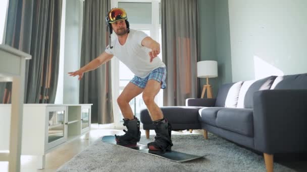 Vídeo divertido. Homem de shorts e uma camiseta retrata snowboard em um tapete em um quarto acolhedor. À espera de um inverno nevado — Vídeo de Stock