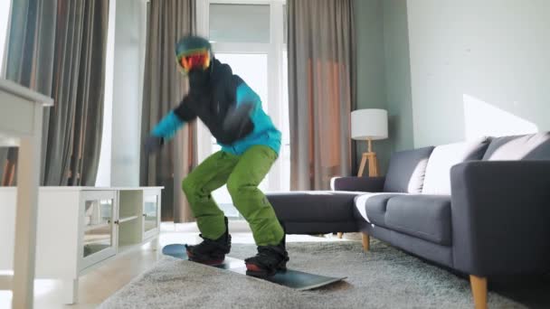Un video divertente. Uomo vestito da snowboarder cavalca uno snowboard su un tappeto in una stanza accogliente. Aspettando un inverno nevoso — Video Stock