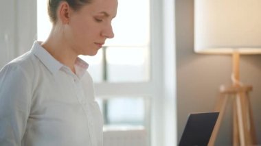 Kadın sıcak bir odada kanepede oturuyor ve gün batımında dizüstü bilgisayarla çalışıyor. Uzak çalışma kavramı.