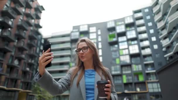 O femeie îmbrăcată formal mergând pe stradă într-un cartier de afaceri cu cafea în mână și vorbind la un apel video pe smartphone — Videoclip de stoc