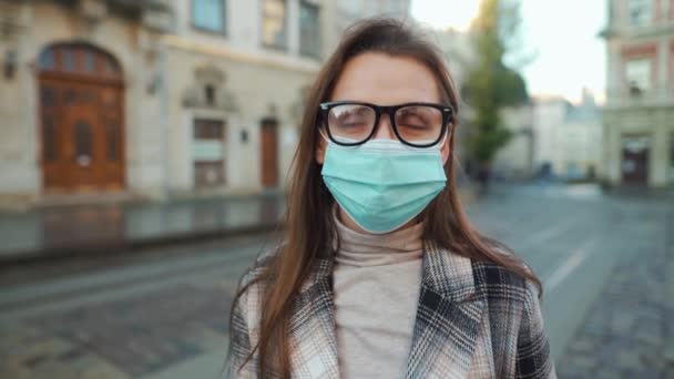 Pandemische bescherming van het Covid-19 coronavirus. Portret van een vrouw in een jas, bril en een beschermend medisch masker. Ze staat midden op het plein.. — Stockvideo