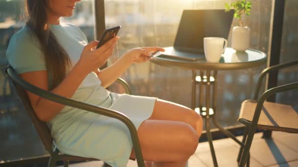 Frau sitzt auf dem Balkon vor dem Hintergrund der untergehenden Sonne und tätigt einen Online-Kauf mit Kreditkarte und Smartphone — Stockvideo