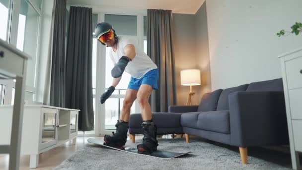 Un video divertente. Uomo in pantaloncini e una T-shirt raffigura snowboard su un tappeto in una stanza accogliente. Aspettando un inverno nevoso — Video Stock