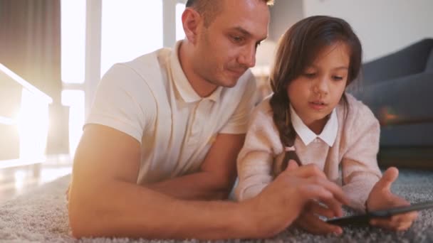 Glücklicher Vater und Tochter genießen die gemeinsame Zeit und nutzen ein Tablet zur Familienunterhaltung, während sie zu Hause im Wohnzimmer auf dem Boden liegen. — Stockvideo