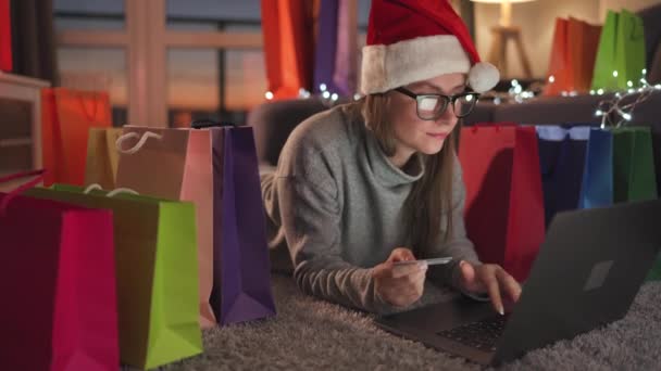 Mujer feliz con gafas con un sombrero de Santa Claus está acostado en la alfombra y hace una compra en línea con una tarjeta de crédito y un ordenador portátil. Bolsas de compras alrededor. — Vídeos de Stock