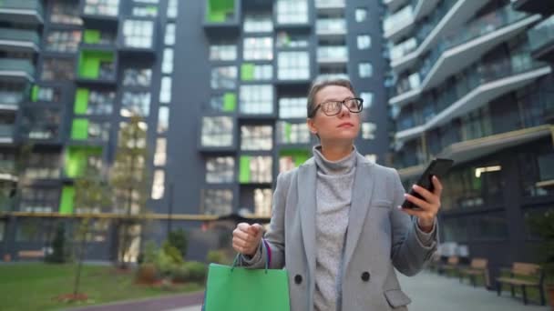 Donna che cammina su un quartiere degli affari con le borse della spesa e utilizzando lo smartphone. Stile di vita urbano e concetto di tecnologia digitale — Video Stock