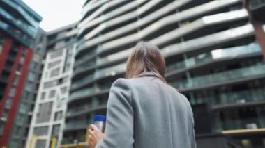 Resmi olarak giyinmiş bir kadın elinde termo bardakla bir iş bölgesinde duruyor ve akıllı telefon kullanıyor. Kamera onun etrafında dönüyor.