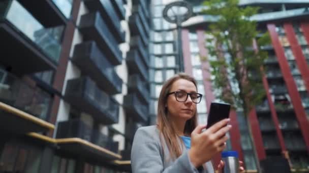 O femeie îmbrăcată formal mergând pe stradă într-un cartier de afaceri cu cafea în mână și folosind un smartphone — Videoclip de stoc