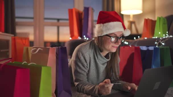 Mujer feliz con gafas con un sombrero de Santa Claus está acostado en la alfombra y hace una compra en línea con una tarjeta de crédito y un ordenador portátil. Bolsas de compras alrededor. — Vídeos de Stock
