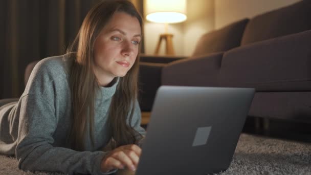 Femme est allongée sur le tapis et travaille sur un ordinateur portable le soir. Concept de télétravail. — Video