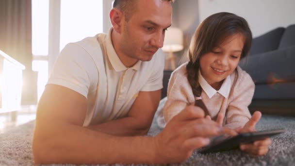 Feliz padre e hija disfrutando del tiempo juntos y utilizando una tableta para el entretenimiento familiar mientras están tumbados en el suelo en la sala de estar en casa. — Vídeo de stock