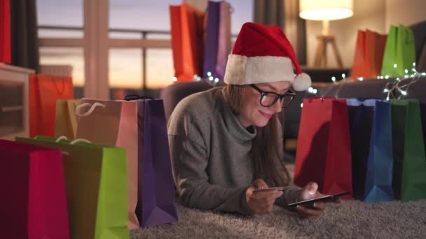 Donna felice con gli occhiali indossa un cappello di Babbo Natale è sdraiato sul tappeto e fa un acquisto online con carta di credito e smartphone. Shopping bag in giro. — Video Stock