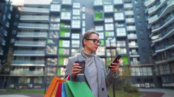 Kobieta stoi w dzielnicy biznesowej, trzyma kawę na wynos, torby na zakupy i używa smartfona. Koncepcja miejskiego stylu życia i technologii cyfrowych — Wideo stockowe