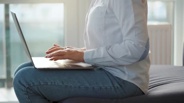 Mujer está sentada en el sofá en una habitación acogedora y trabajando en un ordenador portátil al atardecer. Concepto de trabajo remoto. — Vídeo de stock