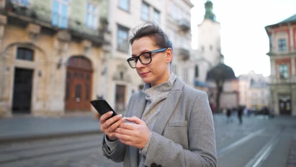 Donna con gli occhiali che indossa un cappotto camminando lungo una vecchia strada e utilizzando lo smartphone. Rallentatore — Video Stock