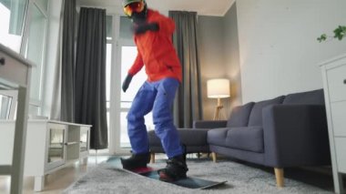 Eğlenceli video. Snowboard 'cu gibi giyinmiş bir adam rahat bir odada halının üstünde snowboard sürüyor. Karlı bir kış bekliyor