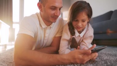 Baba ve kızı birlikte vakit geçiriyorlar ve evde oturma odasında yerde yatarken aile eğlencesi için tablet kullanıyorlar..