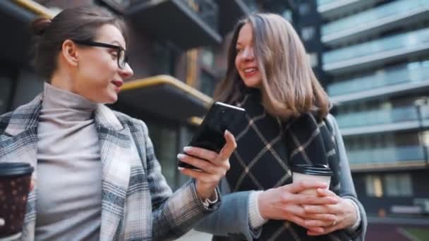 Dos mujeres felices caminando con café para llevar y hablando con interés entre ellas en el distrito de negocios. Movimiento lento — Vídeo de stock